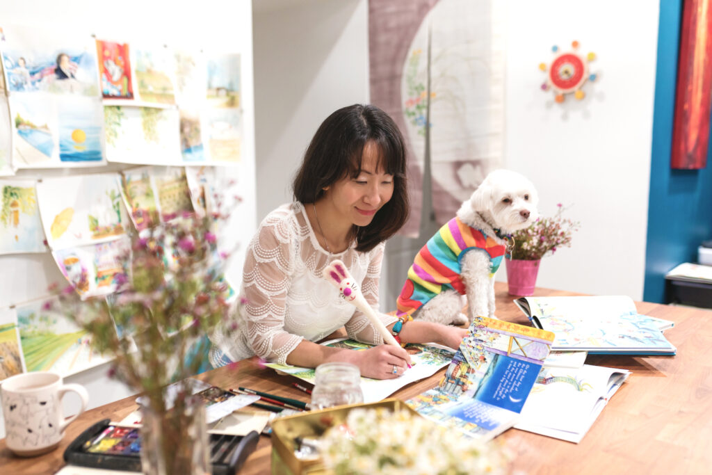Illustrator Alina Chau sitting at her desk in her studio, with pages of her art on the wall behind her and on the table. A small white dog wearing a multicolored striped coat sits on the table beside her.